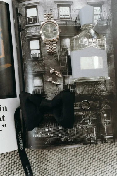 Groom accessories flatlay with gold wristwatch, cologne bottle, cufflinks, and black bow tie on textured fabric over city photo print