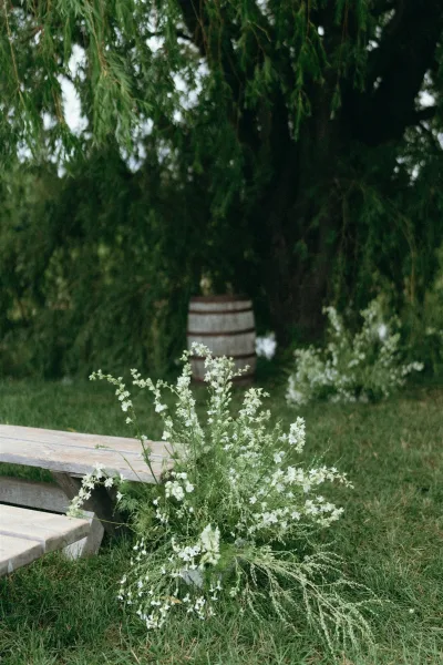 Ceremony aisle florals with white wildflowers and greenery beside a wooden bench and wine barrel on a grassy lawn under trees