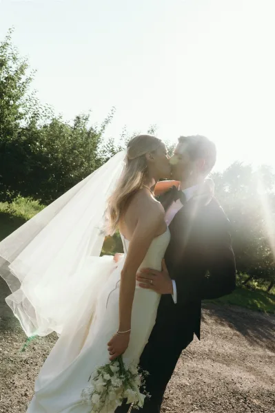 Wedding kiss portrait of bride and groom kissing, backlit by sun flare on a gravel path with trees, her veil lifting as they embrace