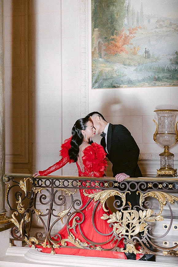 Wedding kiss portrait of bride and groom kissing on a staircase, her dramatic red gown and his black tuxedo by ornate gilded railing