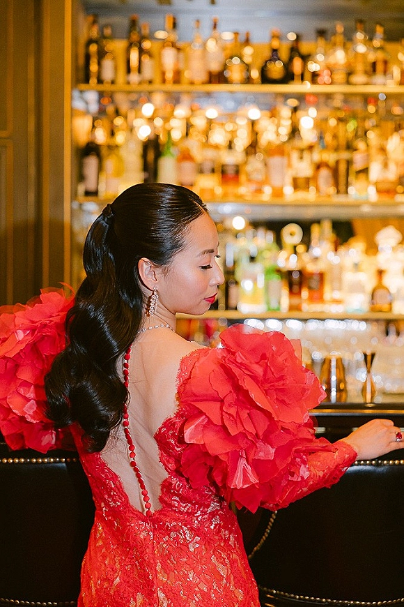 Bridal portrait in a red wedding dress with ruffled sleeves and open back, bride looking over shoulder at a warm, backlit bar shelves
