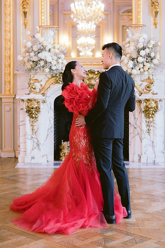 Couple portrait of bride and groom embrace, her red ruffle gown with long train beside a chandelier and marble fireplace in ornate ballroom