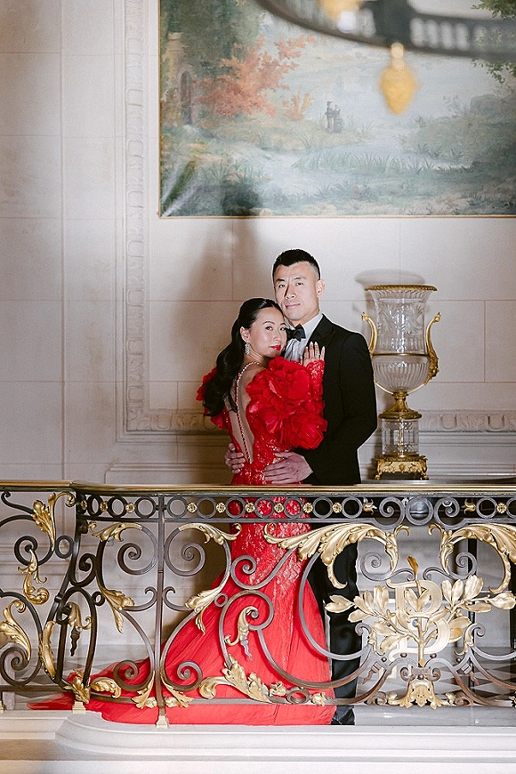 Couple portrait of bride in a red evening gown and groom in black tuxedo with bow tie by an ornate balcony railing under a chandelier