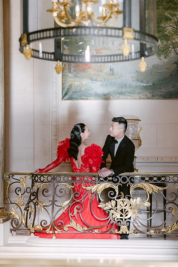 Couple portrait of bride in a dramatic red gown and groom in black tuxedo by an ornate railing beneath a chandelier on a grand staircase landing
