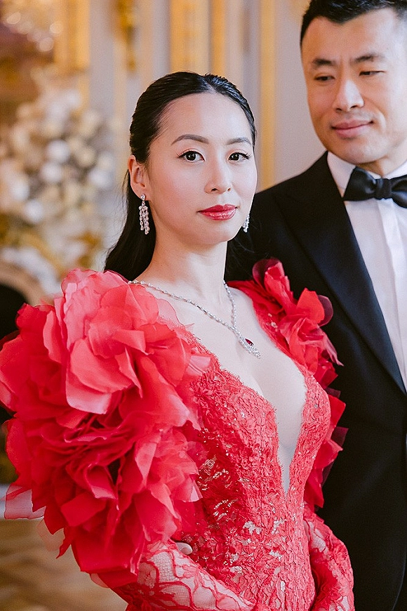 Couple portrait featuring a red wedding dress with ruffled sleeves and lace bodice, groom in tuxedo under warm indoor lighting with blurred florals