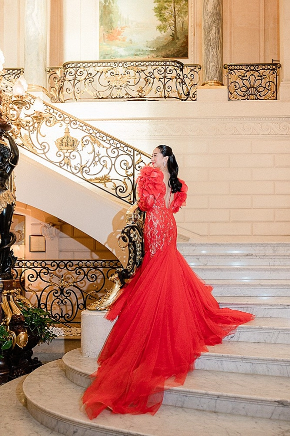Bridal portrait in a red wedding dress with tulle skirt and long train, posed on a grand marble staircase under a chandelier
