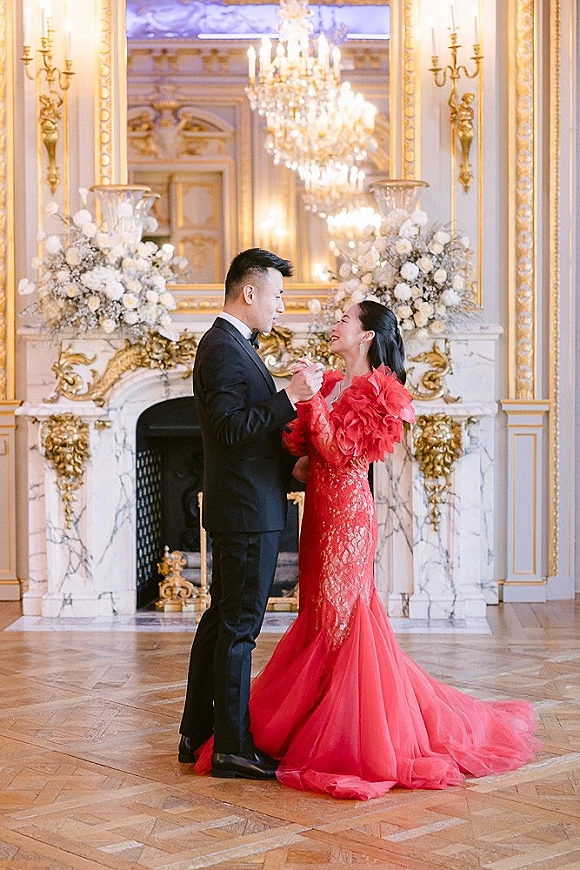 Couple portrait of bride in red ruffled gown and groom in black tuxedo holding hands before an ornate fireplace and gilded mirror