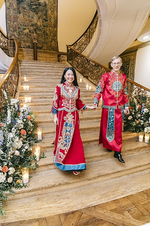 Couple portrait of a red qipao bride and groom tang suit holding hands on a grand staircase with gold embroidery, candles and florals