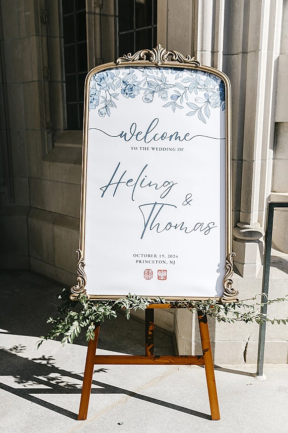 Wedding welcome sign in a gold frame on an easel with floral illustration and calligraphy, set by a sunlit stone building walkway