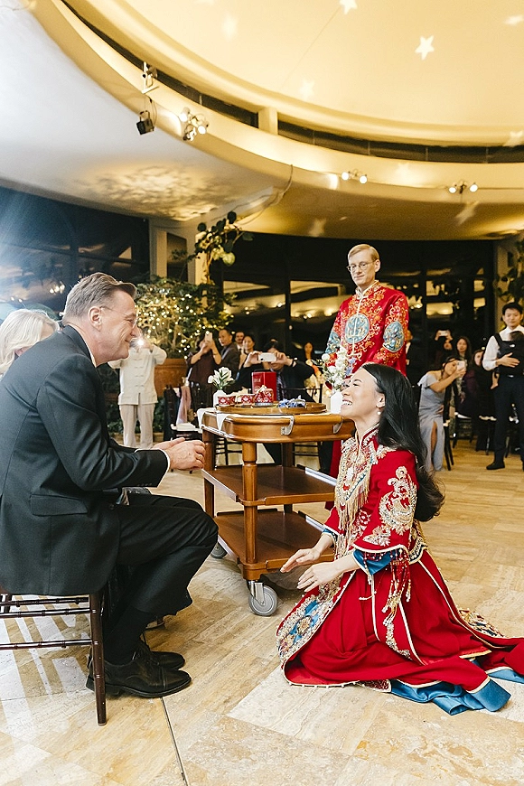 Wedding tea ceremony as bride in a red embroidered dress and groom in black suit serve tea to seated parents in an indoor reception space