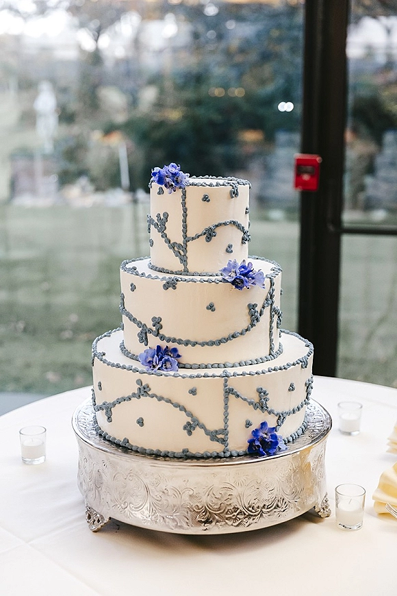 Wedding cake with blue piping on a silver stand, three tiers with sugar flowers and votive candles by a window wall with garden bokeh lights