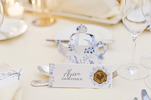 Wedding place card with wax seal place card detail, calligraphy and floral illustration beside blue-and-white teapot, glasses, and flatware on white linen table