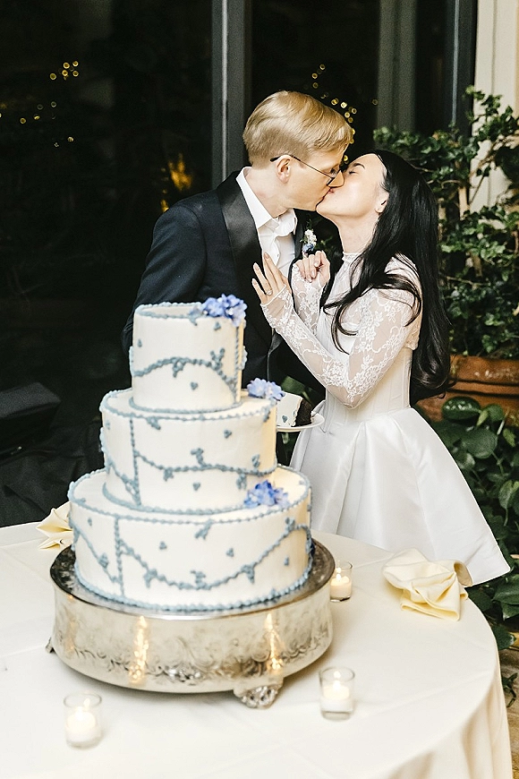 Wedding kiss beside a three-tier white and blue cake on a stand, bride in long sleeve lace dress and groom in tux, greenery wall behind