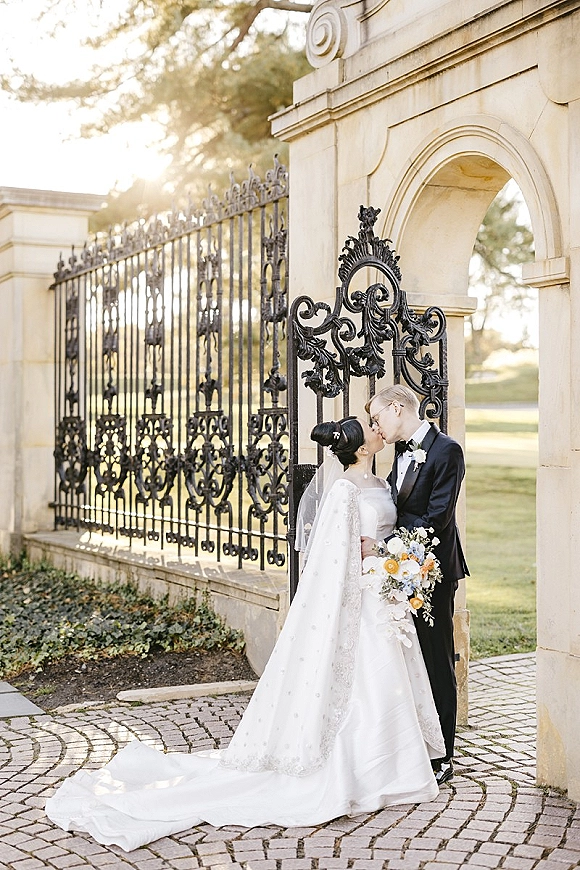 Wedding kiss portrait of bride and groom kissing, her cathedral veil and bouquet against a wrought iron gate and stone archway backdrop
