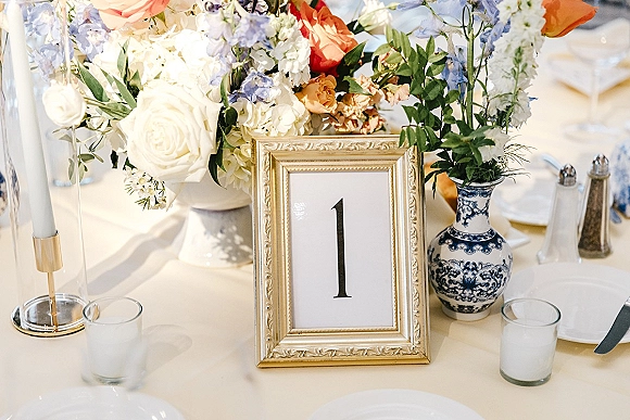 Wedding table number in a gold frame beside a blue and white porcelain vase centerpiece with roses, candles, and formal place settings in soft daylight