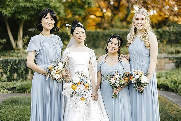 Bridesmaids portrait with bride and bridesmaids in blue dresses holding bouquets, bride in veil and gown, posed in a garden by a stone wall
