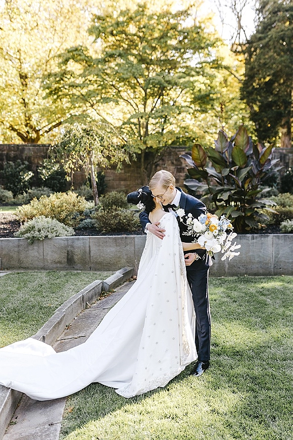 Wedding couple portrait of bride and groom kiss, her long train and veil flowing as she holds a bouquet in a garden by a stone wall