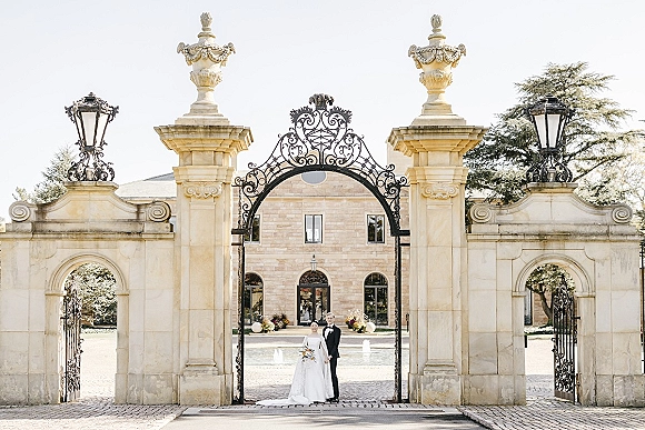 Couple portrait of bride in a strapless wedding dress holding a simple bouquet beside groom in tux at an estate gate entry courtyard