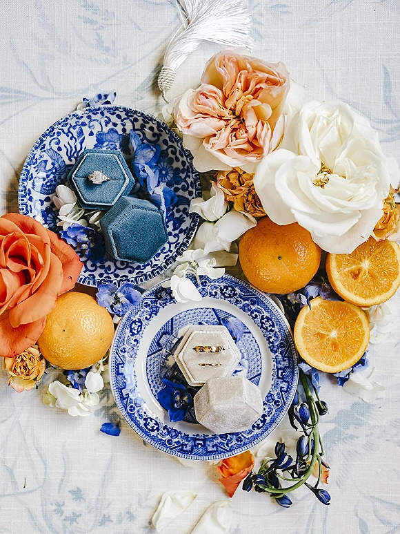 Wedding rings in a ring flat lay with engagement ring and bands in blue boxes on blue-and-white china, styled with oranges and flowers on linen