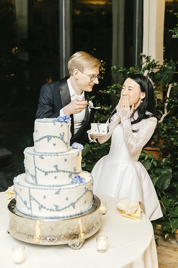 Cake cutting moment as groom feeds bride a bite beside a tiered blue-iced wedding cake on a stand, bride laughing indoors by windows