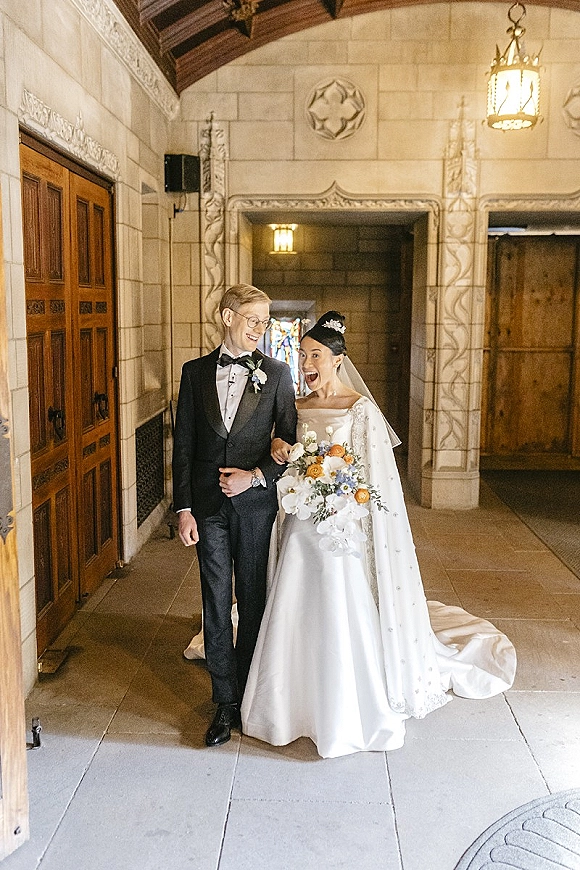 Couple portrait of bride and groom walking arm in arm, bride laughing in tiara and veil, in a stone corridor with stained glass window