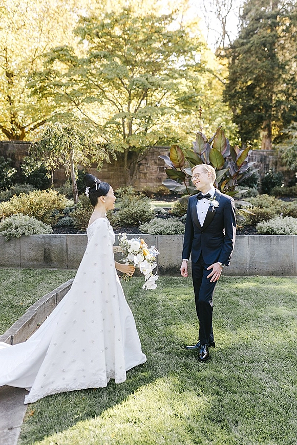 First look moment as the bride in a wedding dress and cape approaches the groom in a tuxedo, holding a white-and-yellow bouquet in a garden lawn