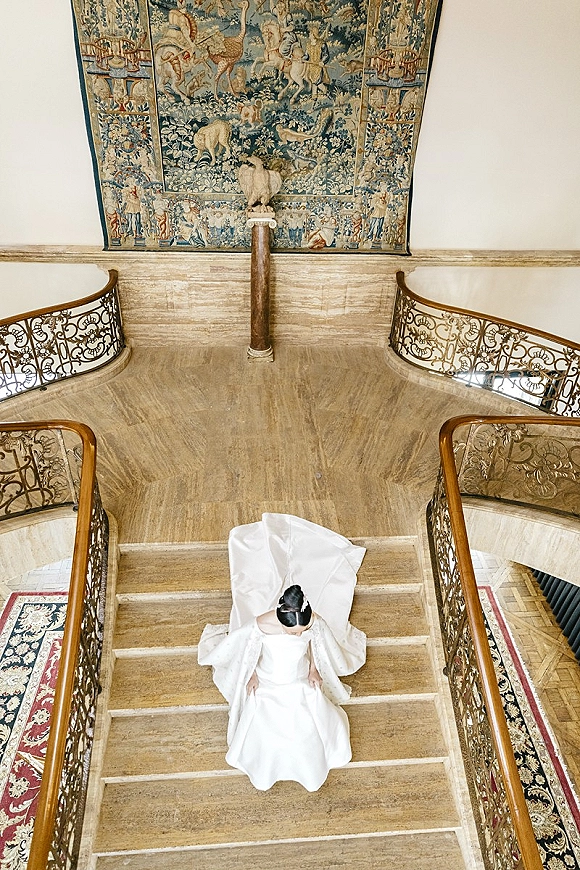 Bridal portrait of a bride on staircase, viewed from above, showing a long satin gown train and veil on a grand marble stairwell