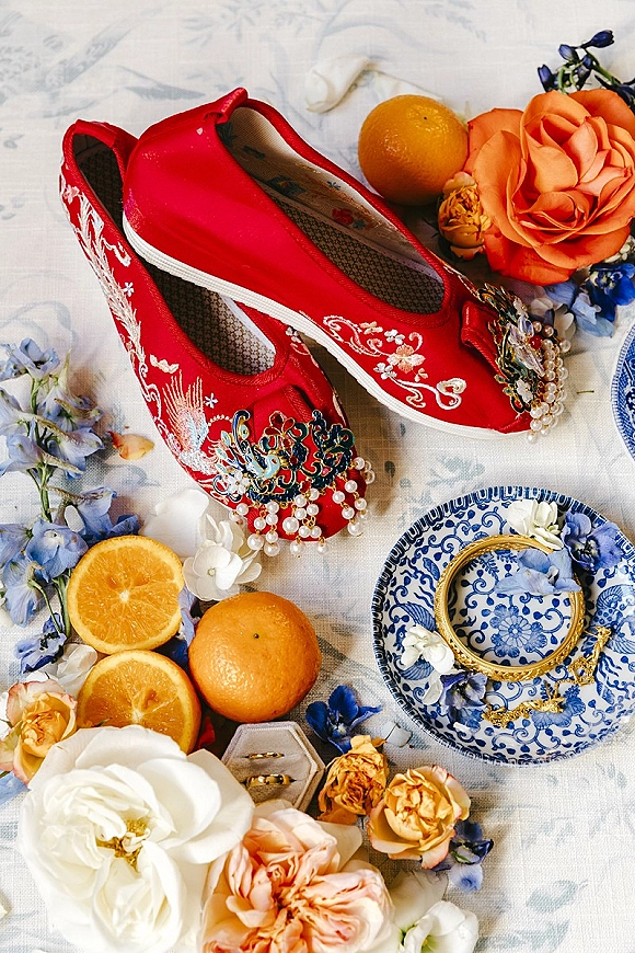 Bridal shoes, red wedding shoes with pearl embellishments and floral clips styled with rings, oranges, and blooms on light fabric surface