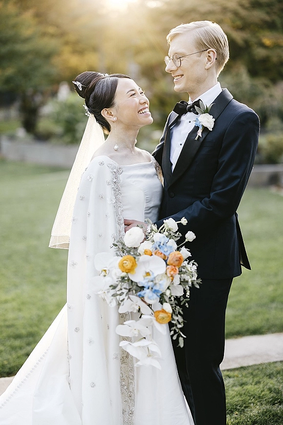 Couple portrait of bride and groom smiling as she looks up at him, her veil and orchid bouquet glowing in sunlit garden light