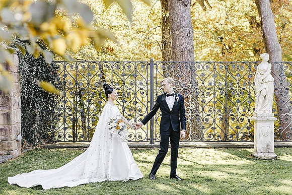 Couple portrait of bride and groom holding hands, her long train and bouquet flowing as they smile by a wrought iron gate and stone wall.