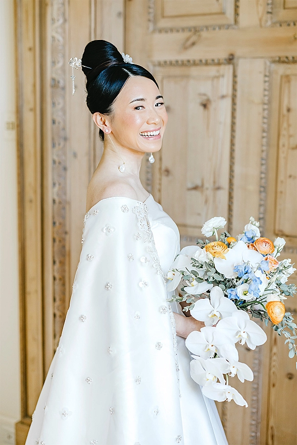 Bridal portrait of a smiling bride in a white cape sleeve gown holding an orchid bouquet, side profile by ornate wooden doors indoors