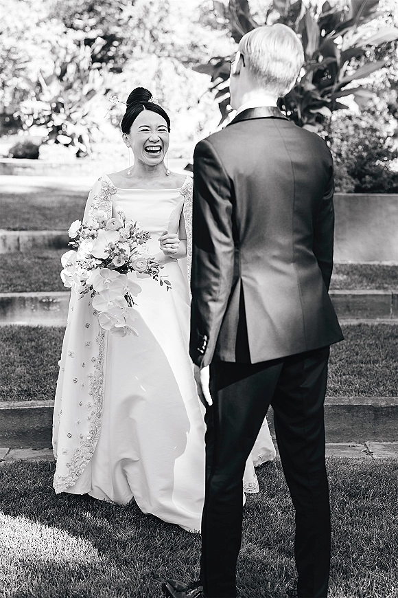 First look moment as bride laughs holding a bouquet, embellished cape and updo with hairpin, facing groom in tuxedo on garden steps