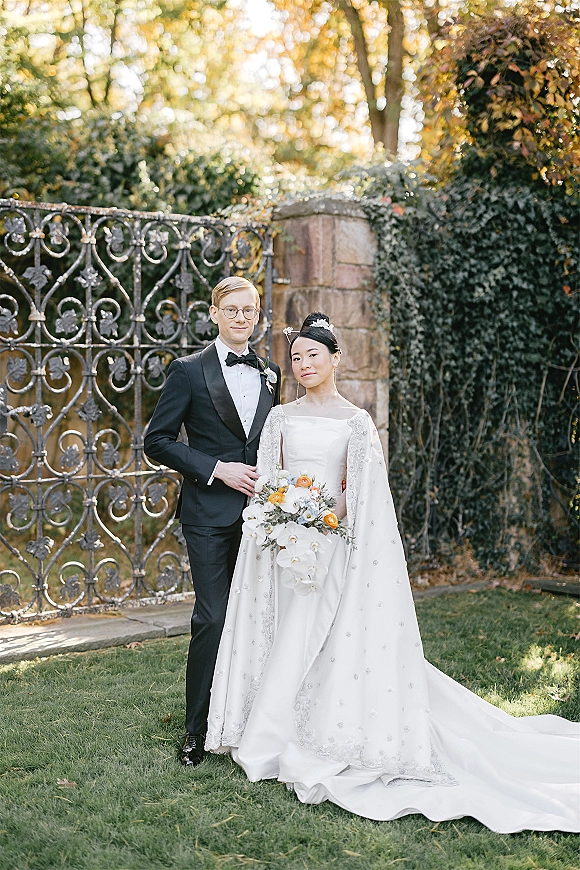 Couple portrait of bride in a flowing cape and long train holding a cascading orchid bouquet beside groom in tux, by an ivy gate