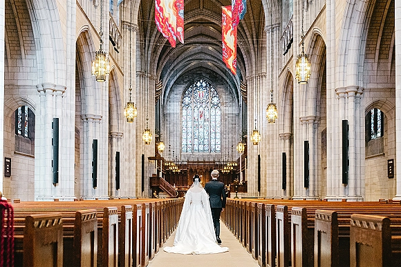 Wedding processional as bride walking down aisle with groom ahead, her cathedral veil and long train flowing past wooden pews in a vaulted cathedral