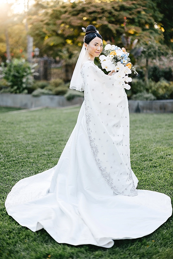 Bridal portrait of a bride holding bouquet with white orchids and blue flowers, wearing a long veil and train on a sunlit garden lawn