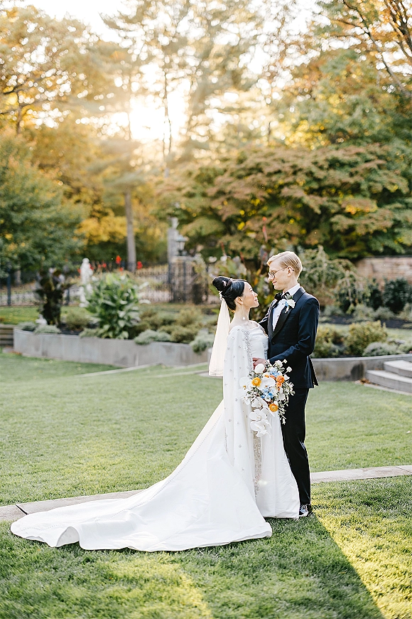 Couple portrait of bride and groom holding each other, her cathedral veil and bouquet flowing in sunset garden light by stone steps