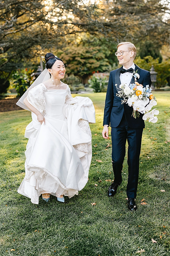Wedding couple portrait of bride and groom walking on a sunlit garden lawn, bride laughing in veil and gown, holding orchid bouquet