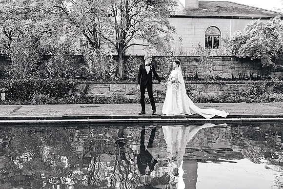 Couple portrait in a black and white wedding portrait, bride and groom holding hands by a reflecting pool, long veil and bouquet visible