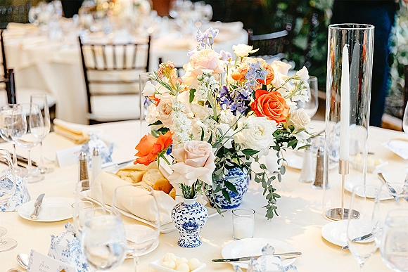 Wedding tablescape with a reception table centerpiece of roses and mixed flowers in blue-and-white porcelain vases, taper candles and place cards on linens
