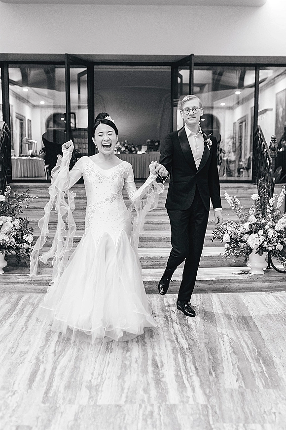 Recessional moment as bride and groom exit holding hands, her tulle-sleeved gown swaying on stone steps by glass doors and urn flowers