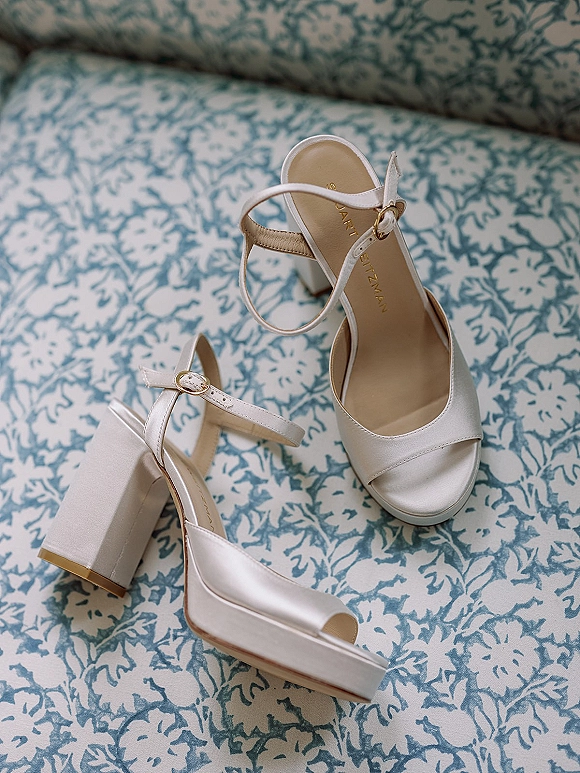 Bridal shoes—white satin wedding heels with ankle straps and gold buckles—rest on a blue and white floral patterned sofa as something blue
