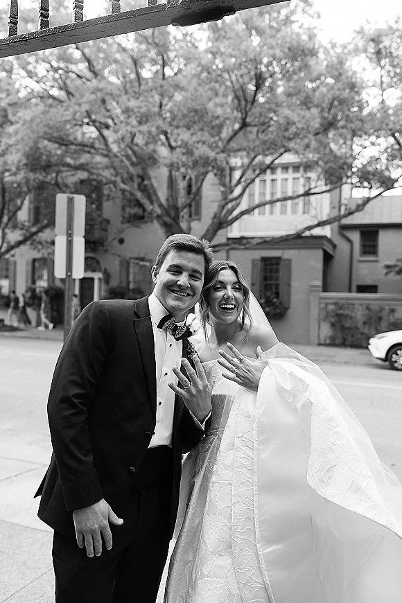 Wedding couple portrait, black and white wedding portrait of bride and groom smiling and showing rings on a city sidewalk with trees and buildings