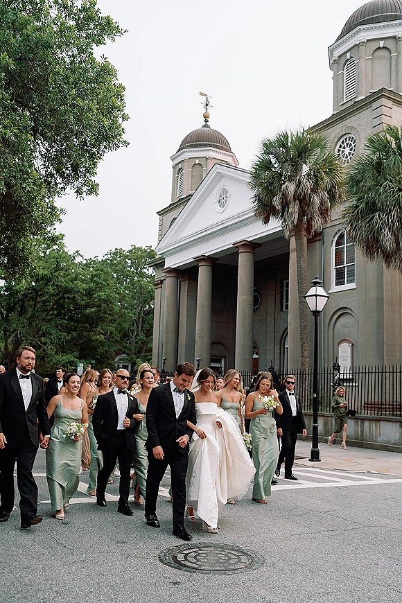 Wedding party exit with bride and groom walking in a crosswalk, bride holding her dress as bridesmaids with bouquets pass a domed church exterior