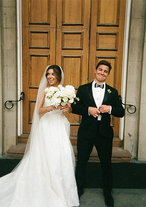 Couple portrait of bride and groom laughing on stone steps, bride holding a white bouquet with veil, groom in tuxedo by wooden doors
