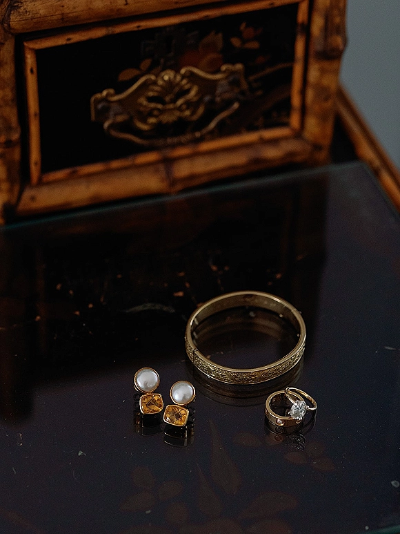 Wedding rings and engagement ring close up on a glass tabletop, engraved gold band beside pearl and gold earrings near a wooden box