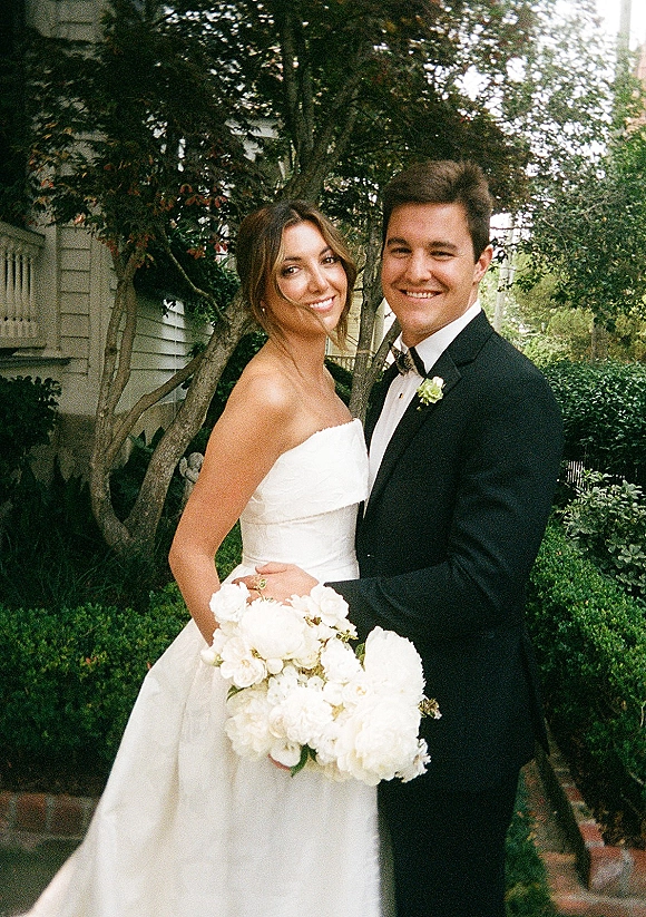 Couple portrait of bride and groom portrait smiling, bride in strapless gown holding white bouquet beside tuxedoed groom in a garden setting