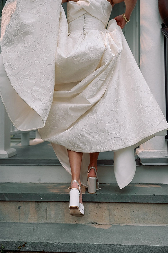 Wedding dress detail of a strapless wedding dress with button-back textured fabric, high-low hem and platform heels on porch steps