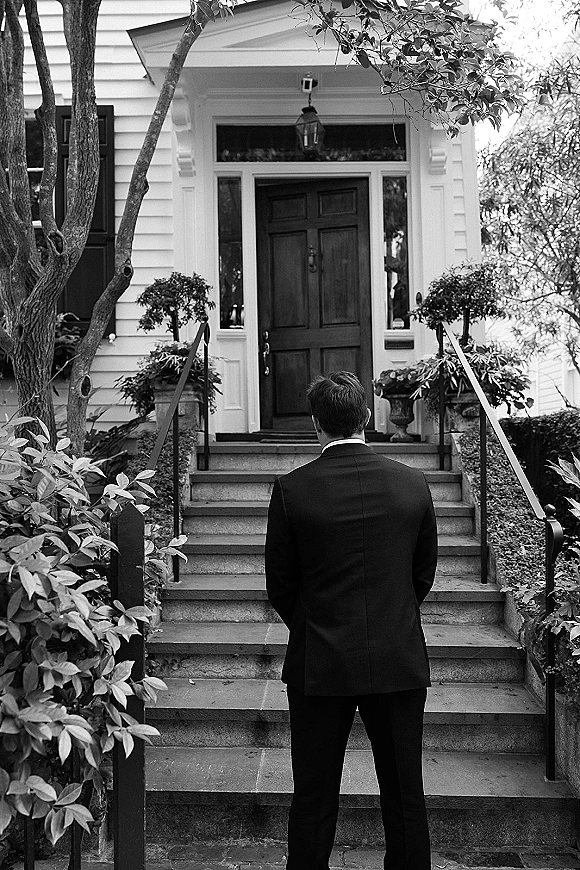 Groom portrait in tuxedo seen from behind, waiting on stone steps by a white house front door with topiary and metal railings.