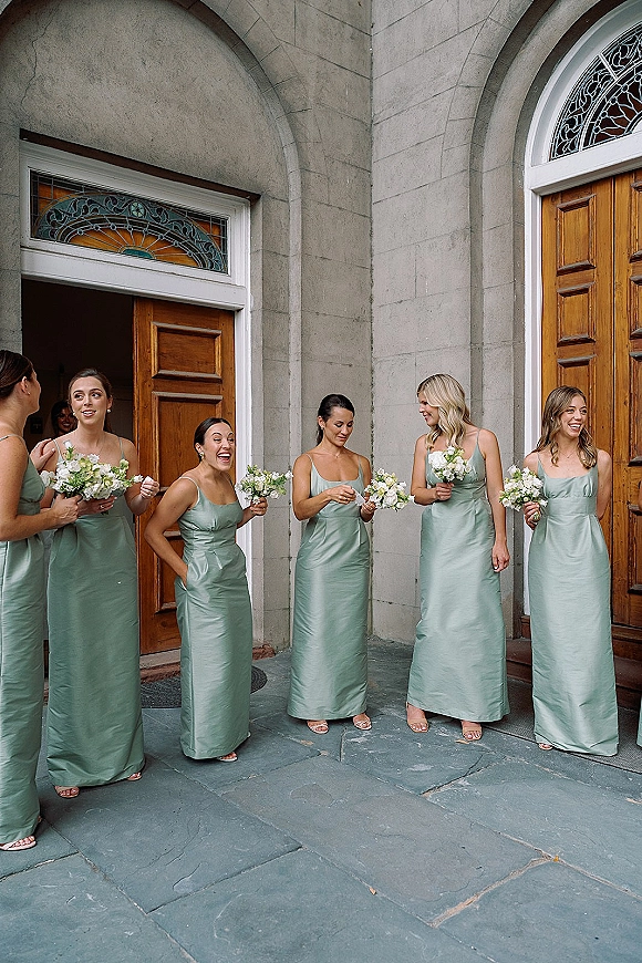 Bridesmaid group in sage green bridesmaid dresses, laughing with bouquets by an arched stone doorway and wooden church doors