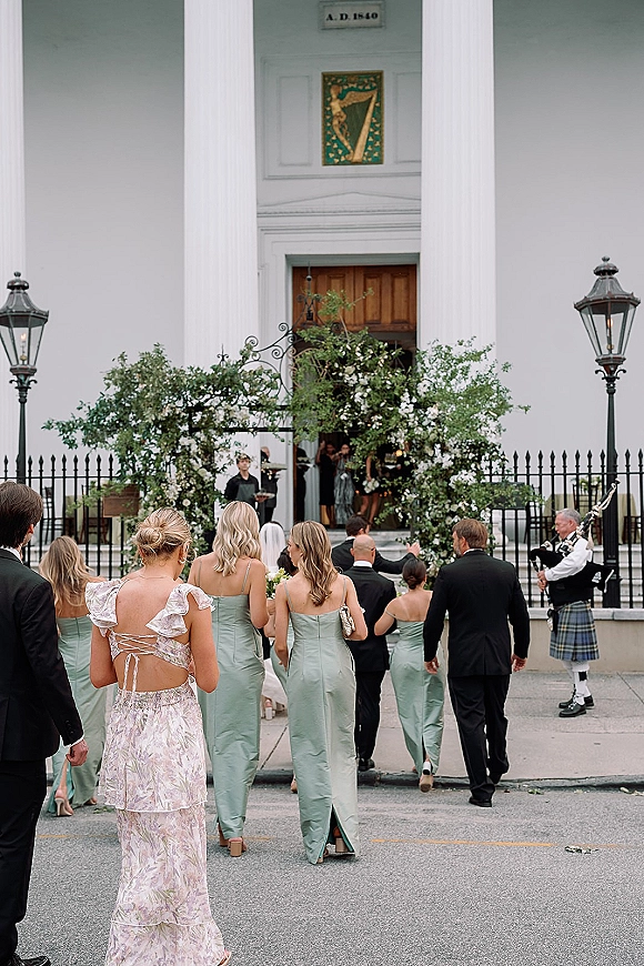 Wedding guests arrival in formal attire walk up steps toward a white-columned venue, passing a greenery arch and bagpiper outdoors
